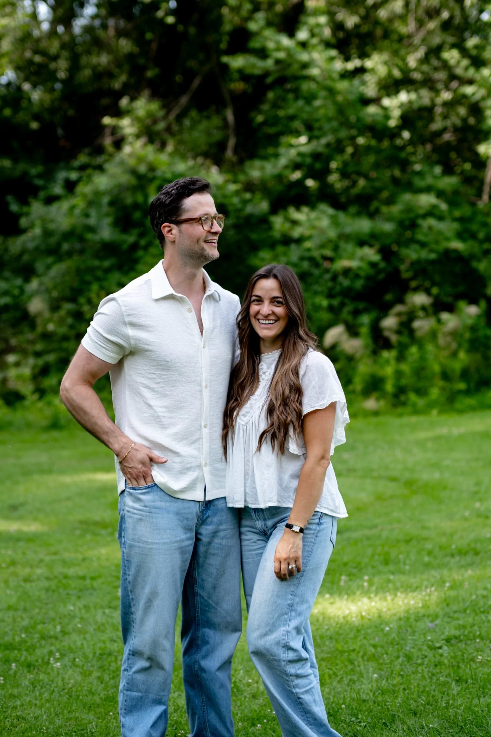 Two people standing in a grassy field with a dense green forest background. The person on the left is a man with dark, curly hair, glasses, wearing a white short-sleeved shirt and blue jeans. The person on the right is a woman with long, brown hair, 