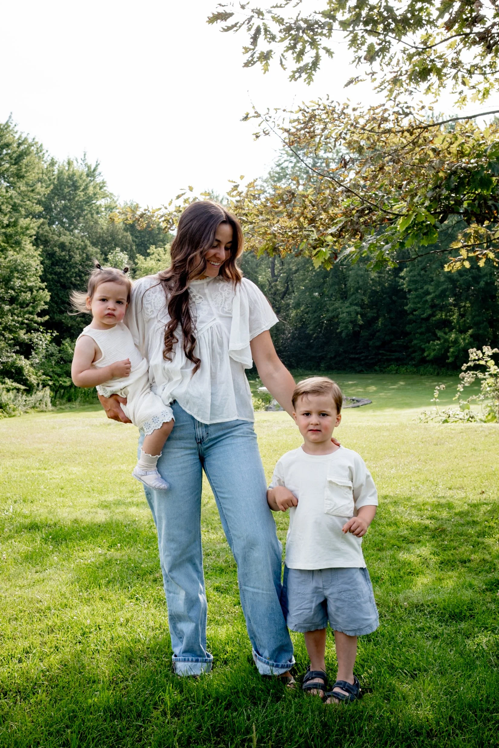 A woman with long, wavy brown hair smiling while holding a young girl with two small pigtails, both wearing white tops, standing outdoors on a grassy field with trees in the background. Two young boys are also present, one being held by the woman and