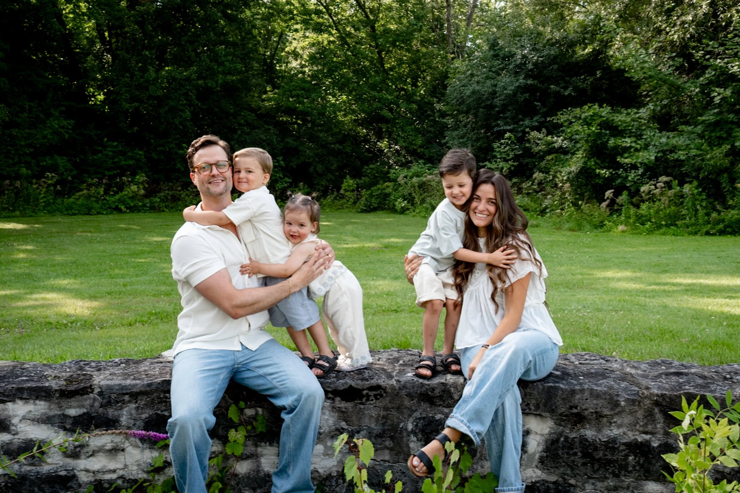 A happy family of two parents and three children outdoors in a park with green trees. The father and two children are sitting on a low stone wall, while the mother and one child are standing behind them, all smiling and embracing.