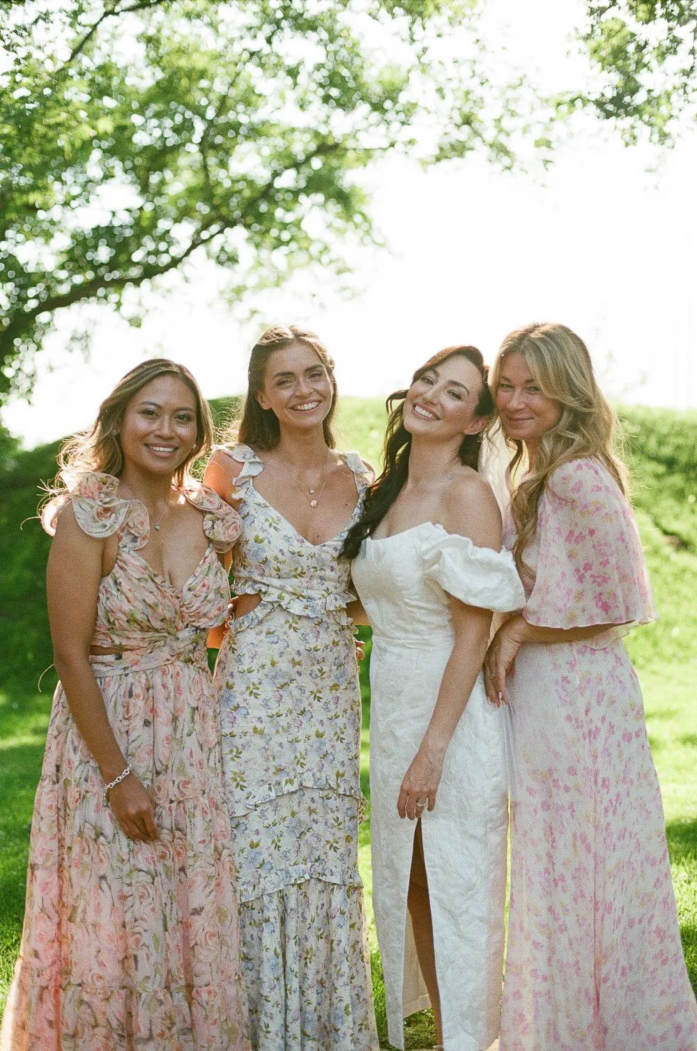 Four women in floral dresses standing outdoors under a tree on a sunny day, smiling at the camera.
