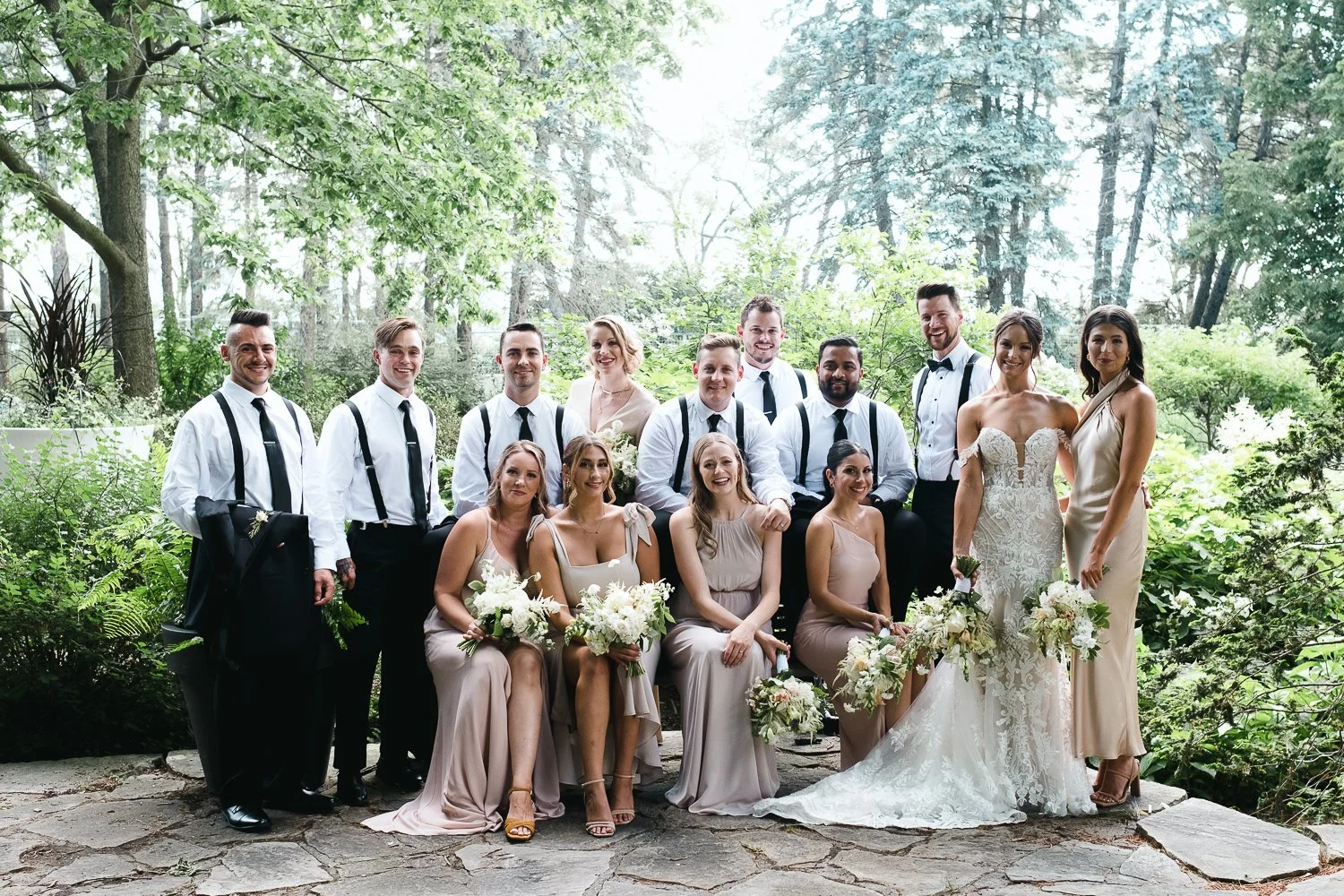 A wedding party outdoors in a green, wooded area with 14 people, including the bride and groom, bridesmaids, and groomsmen, all dressed in formal wedding attire.