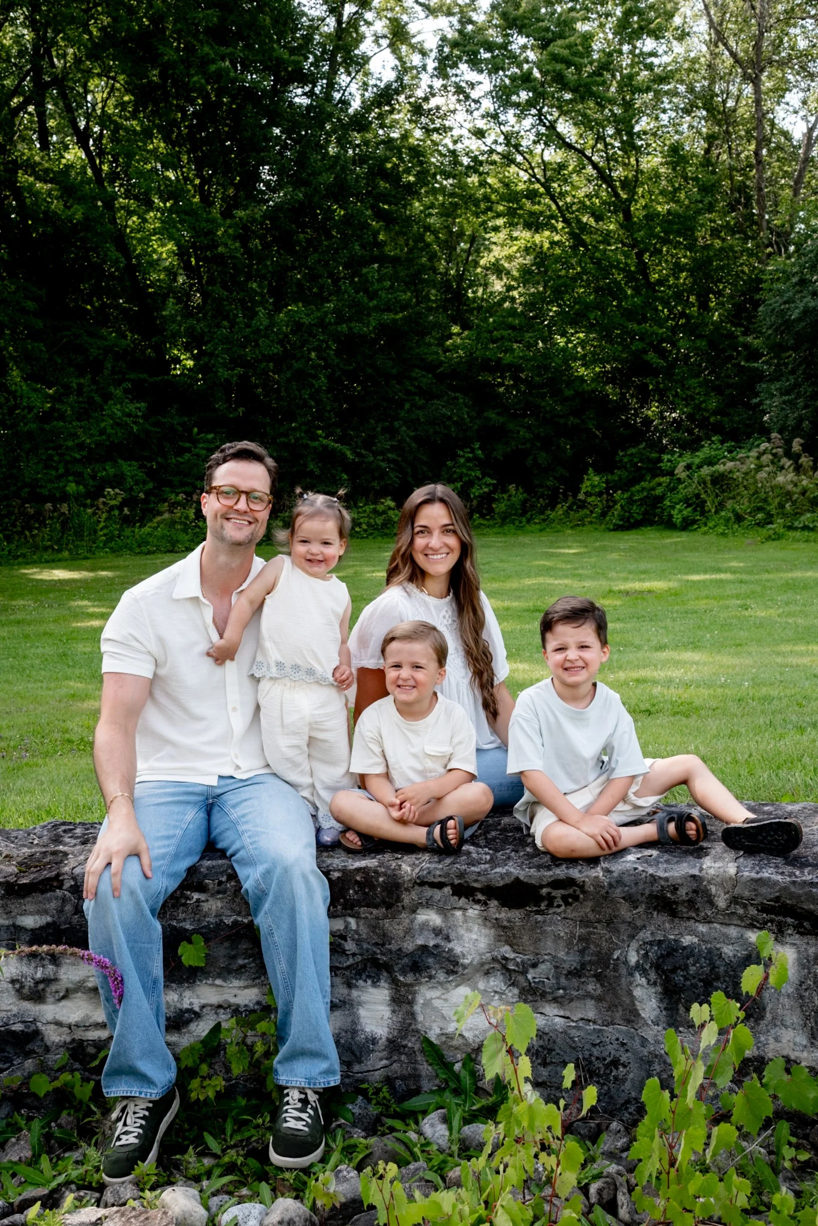 A family of six sitting and standing on a rock in a park, with green grass and tall trees in the background, smiling for the camera.