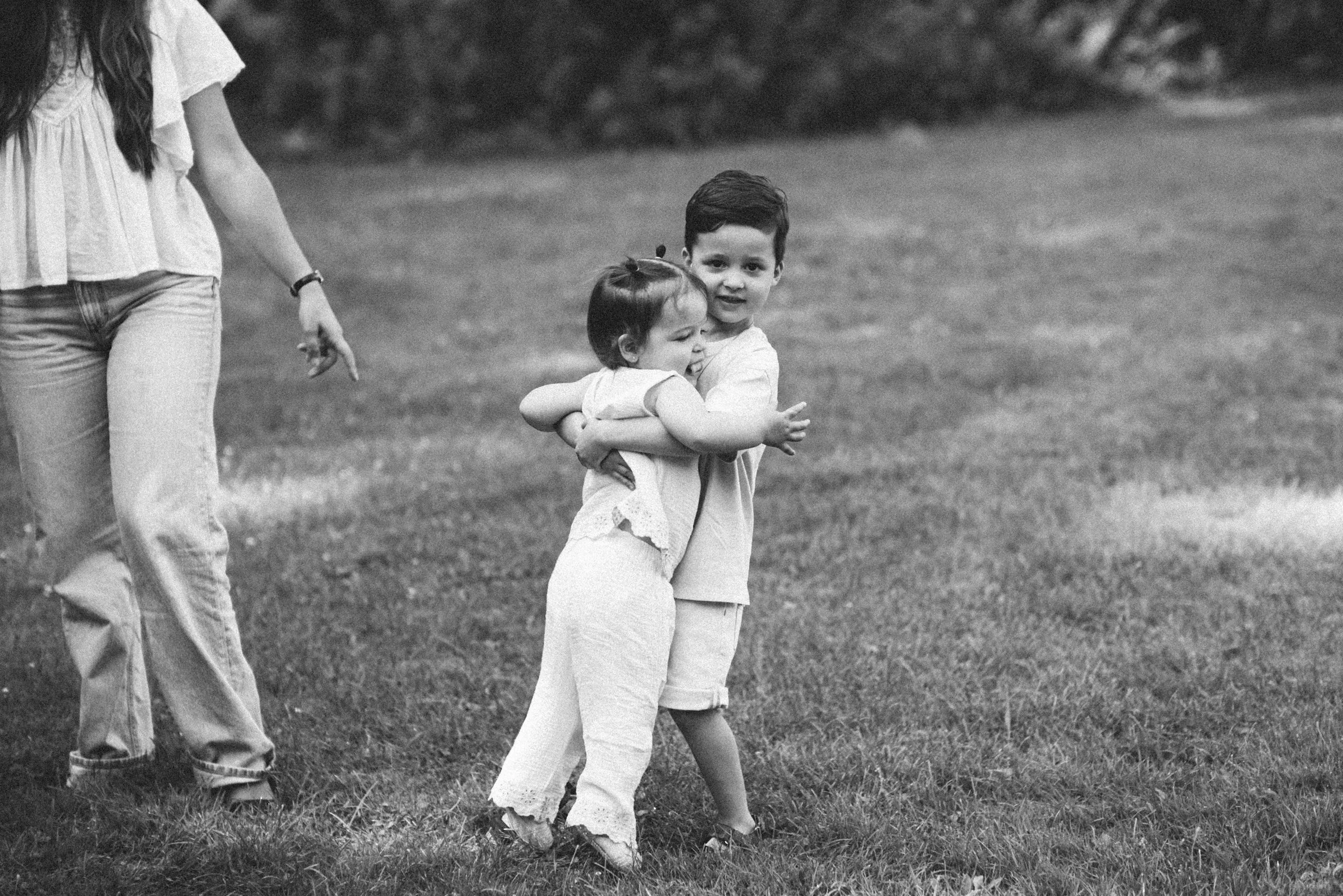 Two young girls hugging each other outdoors on a grassy field, with a woman walking nearby in the background, all in black and white.