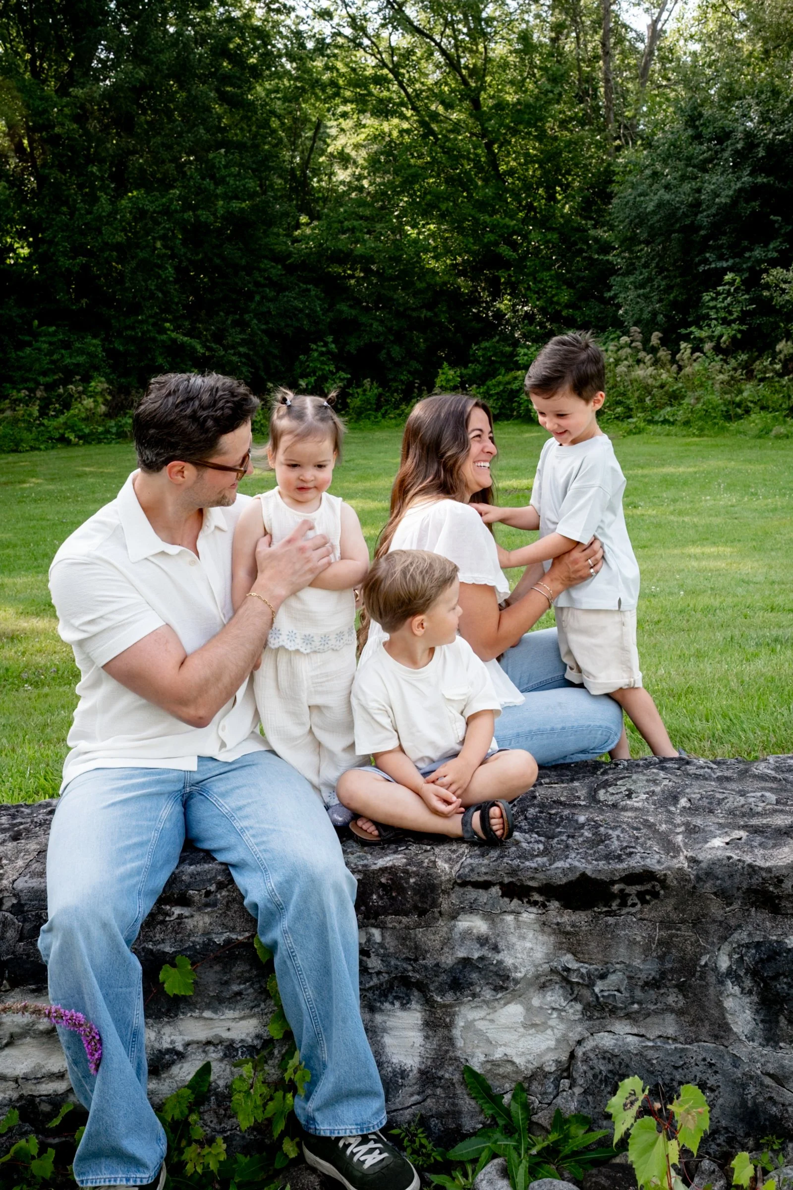 Family of five sitting and standing on a grassy outdoor area with lush green trees in the background. The father is sitting on a rock with two young children, one girl and one boy. The mother is laughing, holding the boy in a white shirt, and two mor