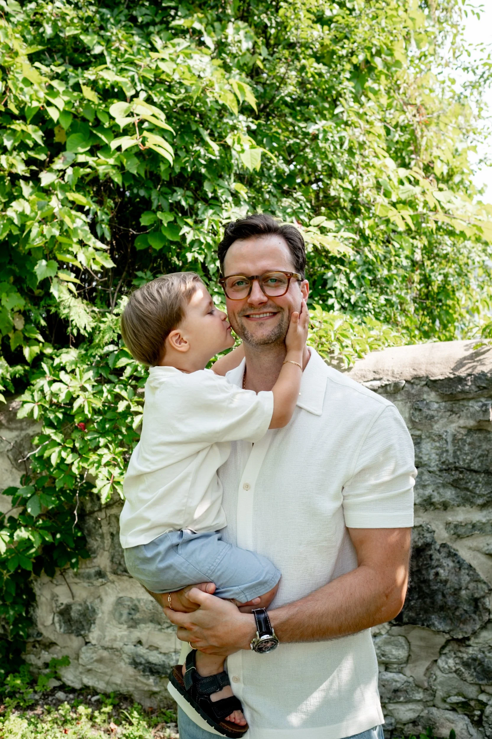 A man is holding a young boy in front of leafy green bushes and a stone wall. The boy is kissing the man on the cheek. Both are smiling, and the man is wearing glasses and a light-colored polo shirt.