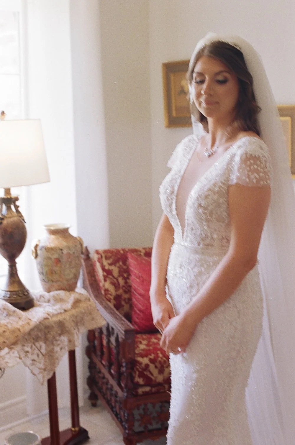 A bride in a white lace wedding dress with a deep V neckline, standing indoors with her eyes closed and hands clasped in front of her, wearing a veil, necklace, and earrings, with vintage furniture and a lamp in the background.