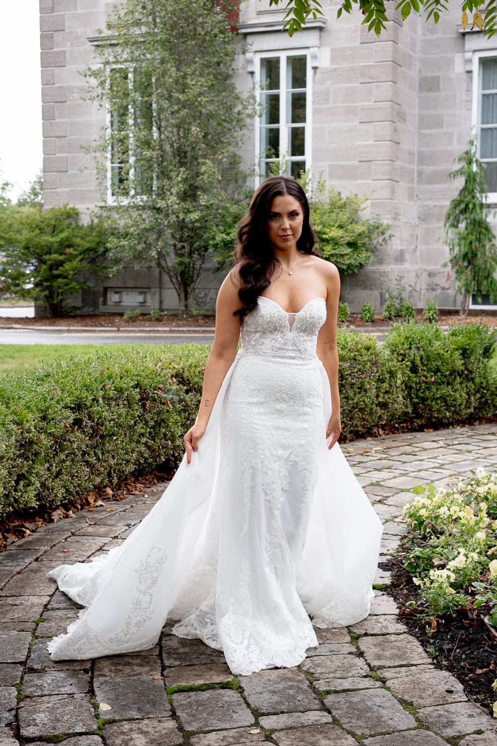 Montreal wedding photographer photographed a woman in a white wedding dress standing outdoors on a cobblestone path, with greenery and a stone building in the background.