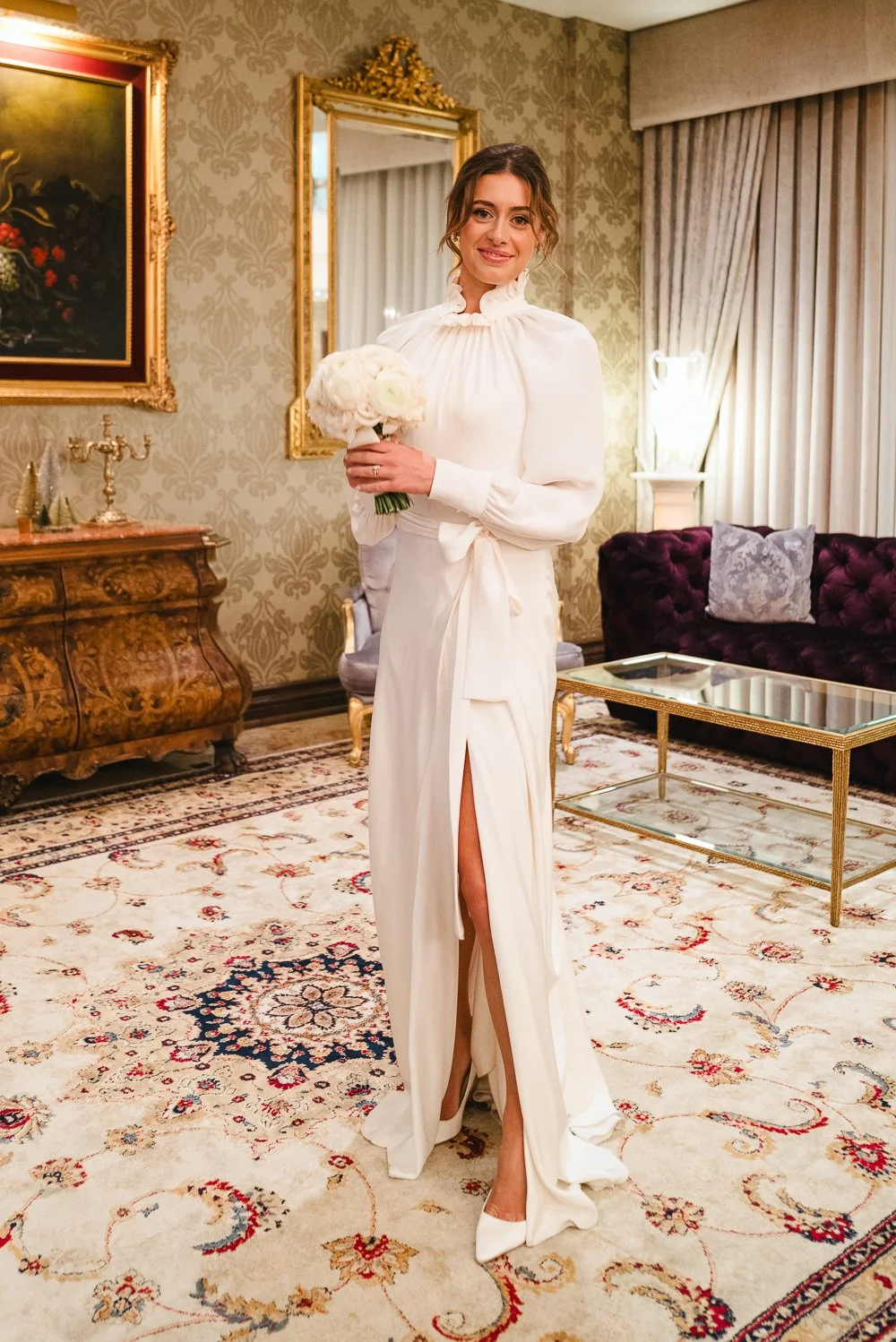 A bride in a white dress holding a bouquet of white roses, standing in a richly decorated room with ornate furniture and patterned wallpaper.