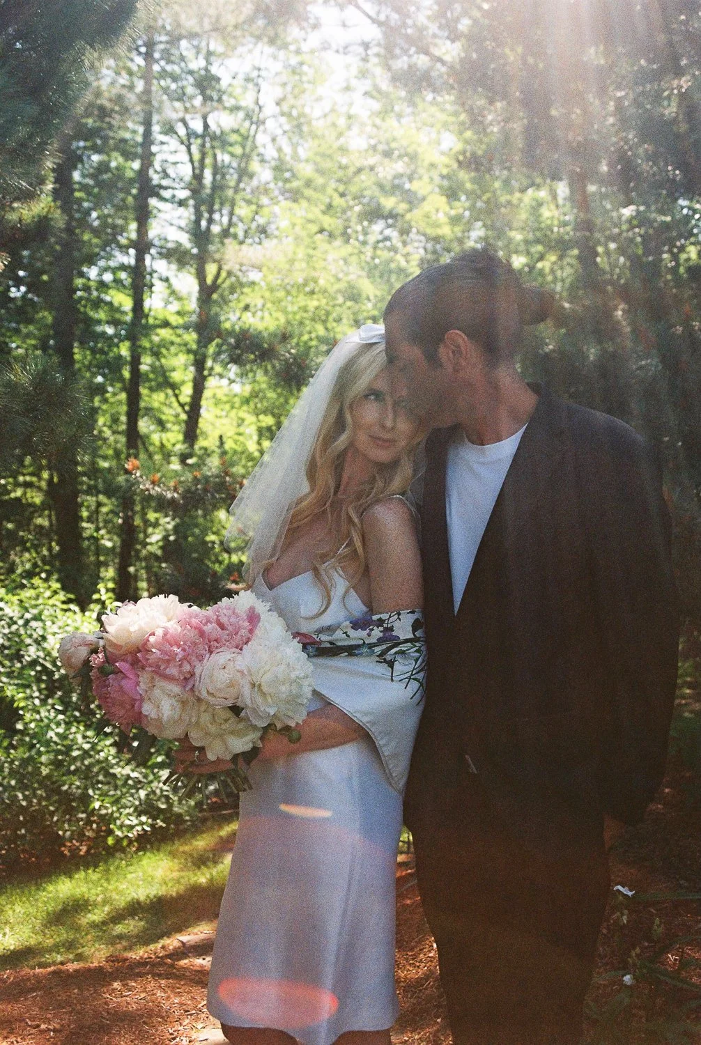 Bride with long blonde hair and white veil holding a bouquet of pink and white flowers, standing close to groom with dark hair, in a forest setting during daytime.