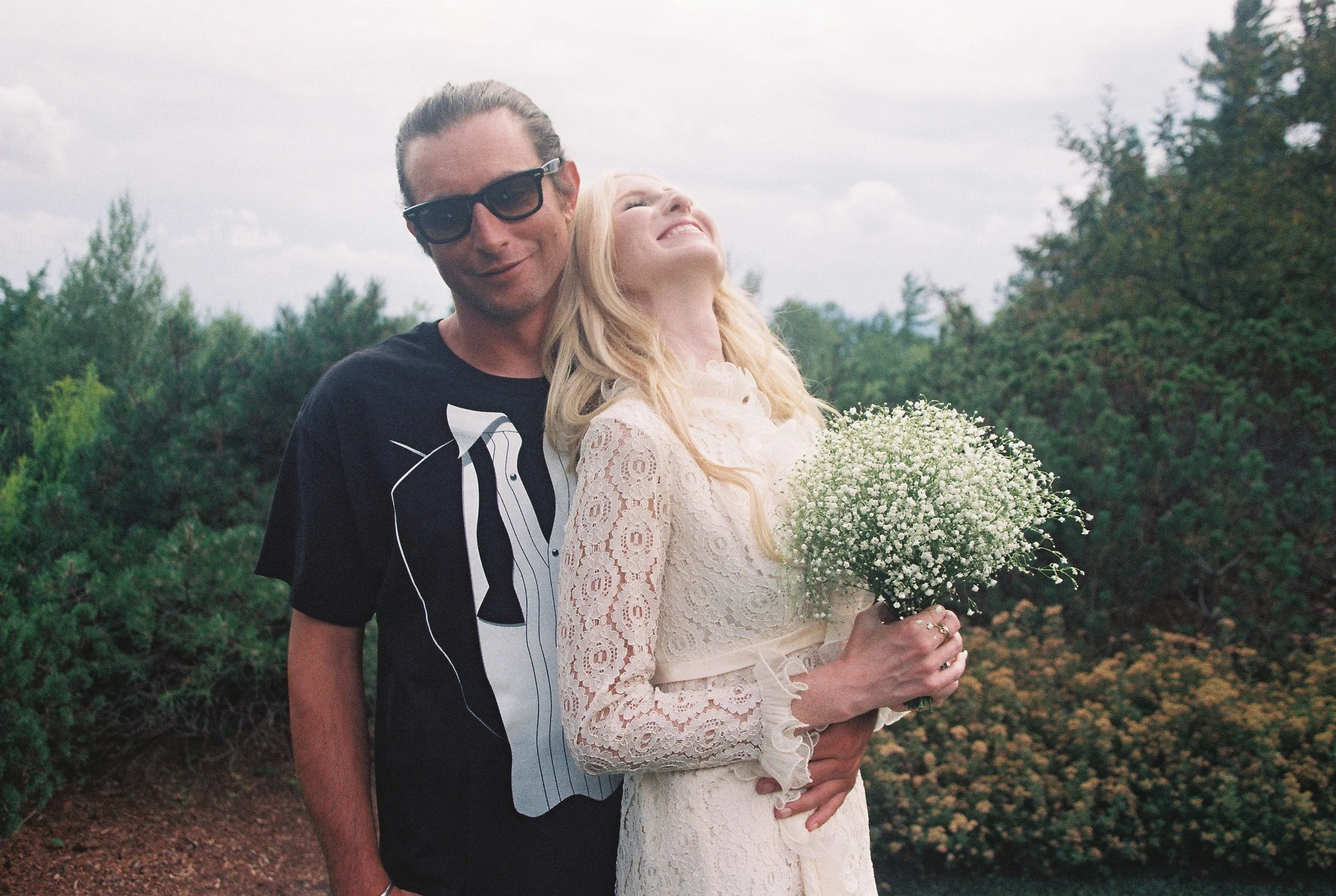 A man with sunglasses and a woman in a white lace dress, holding a bouquet of baby's breath flowers, standing outdoors with trees and a cloudy sky in the background, smiling and enjoying a moment together.
