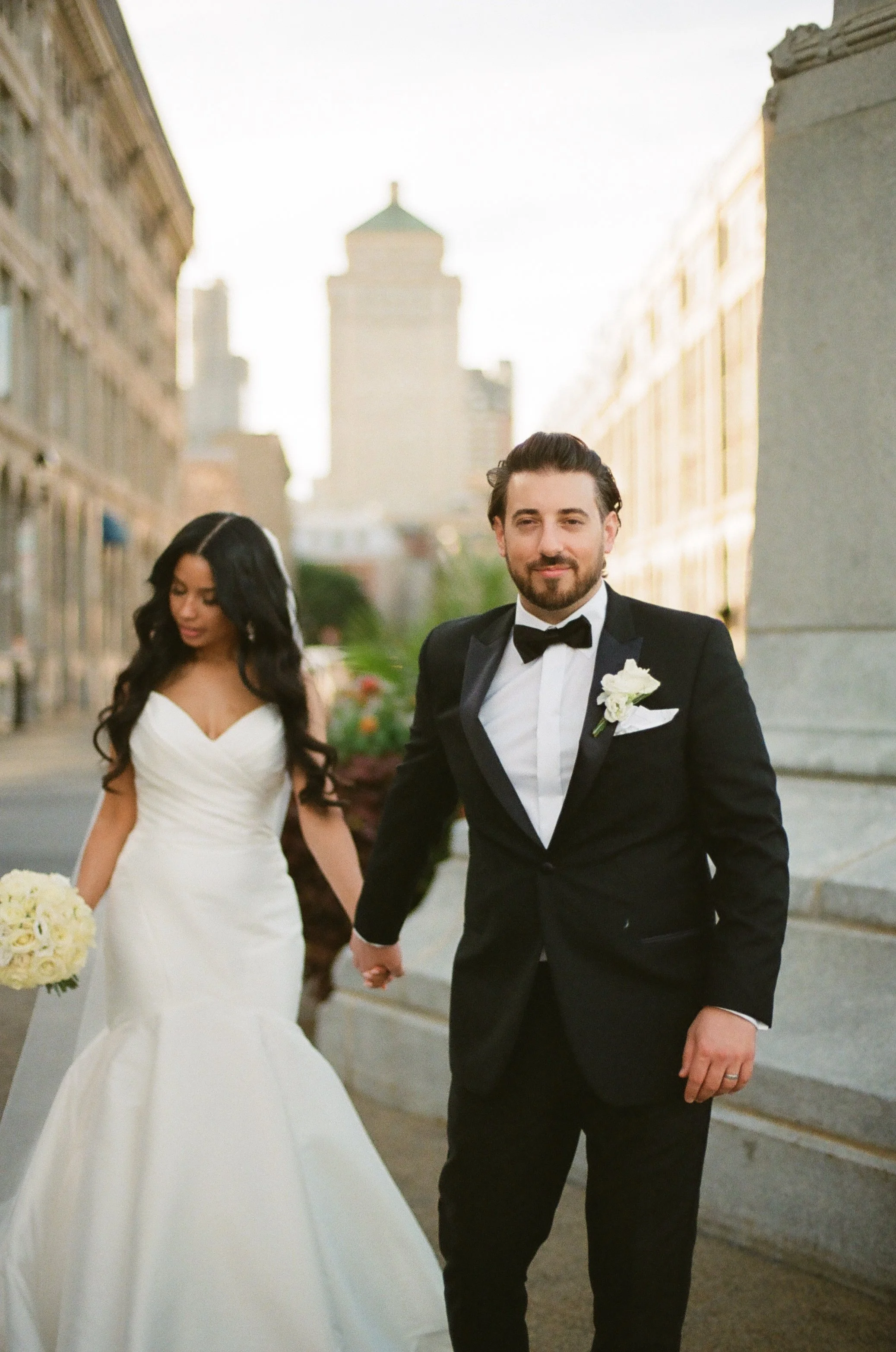 A bride and groom holding hands on a city street during their wedding. The bride is wearing a white wedding gown and the groom is dressed in a black tuxedo with a bow tie.