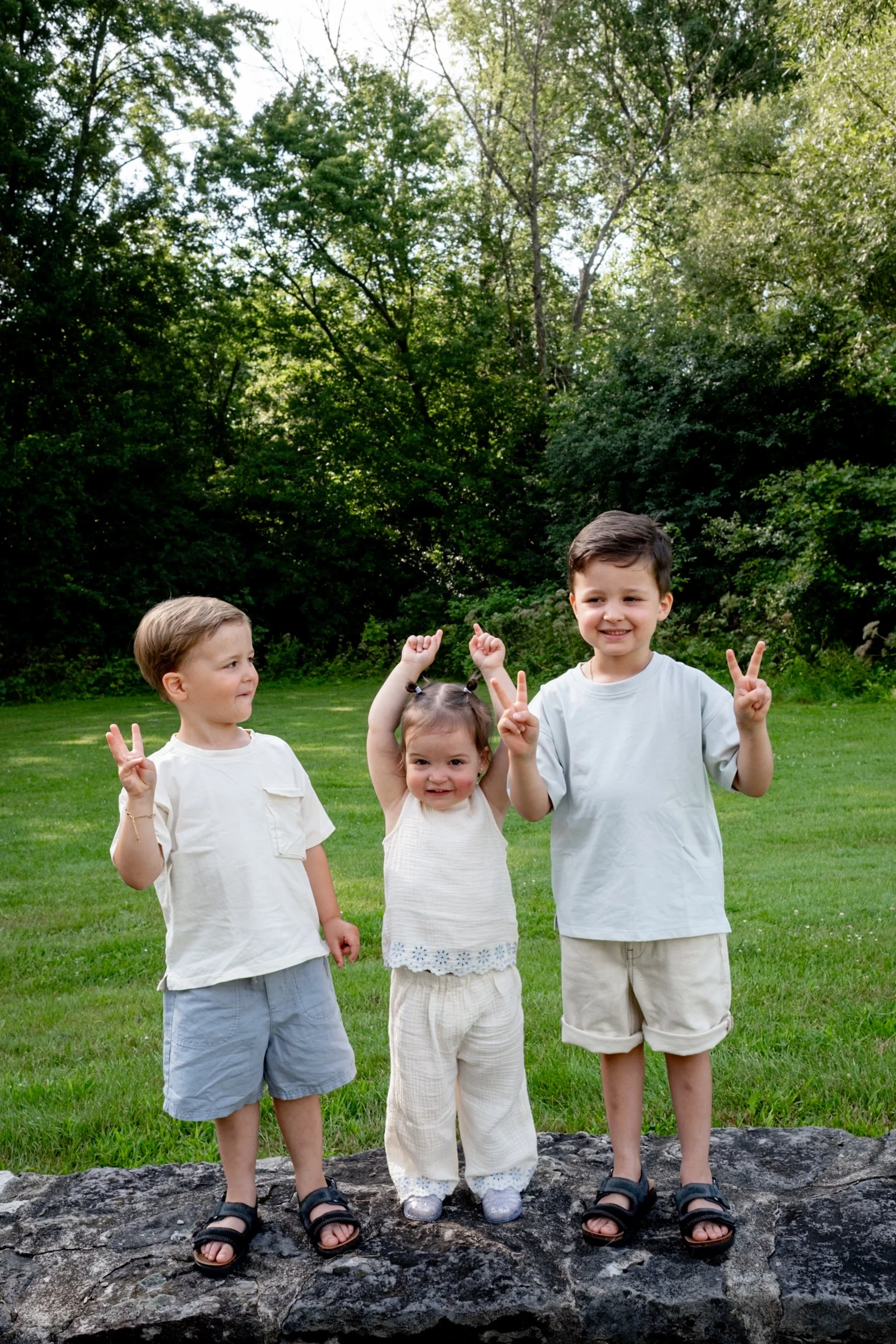 Four children, three boys and one girl, standing on a rock in a park with green grass and trees in the background. They are smiling and making peace signs with their hands.