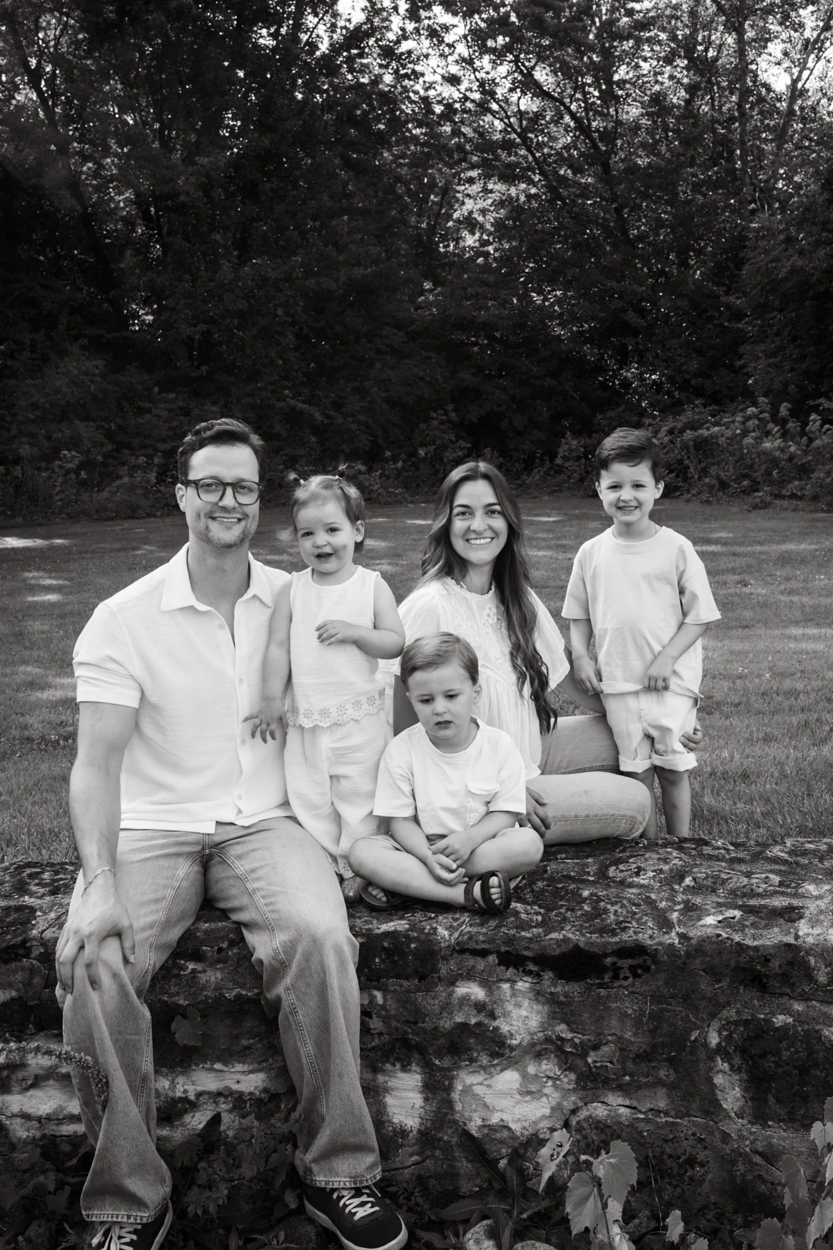 A black and white family portrait of five people outdoors, with trees in the background. The group includes a man, a woman, two boys, and a girl, all smiling and dressed casually.