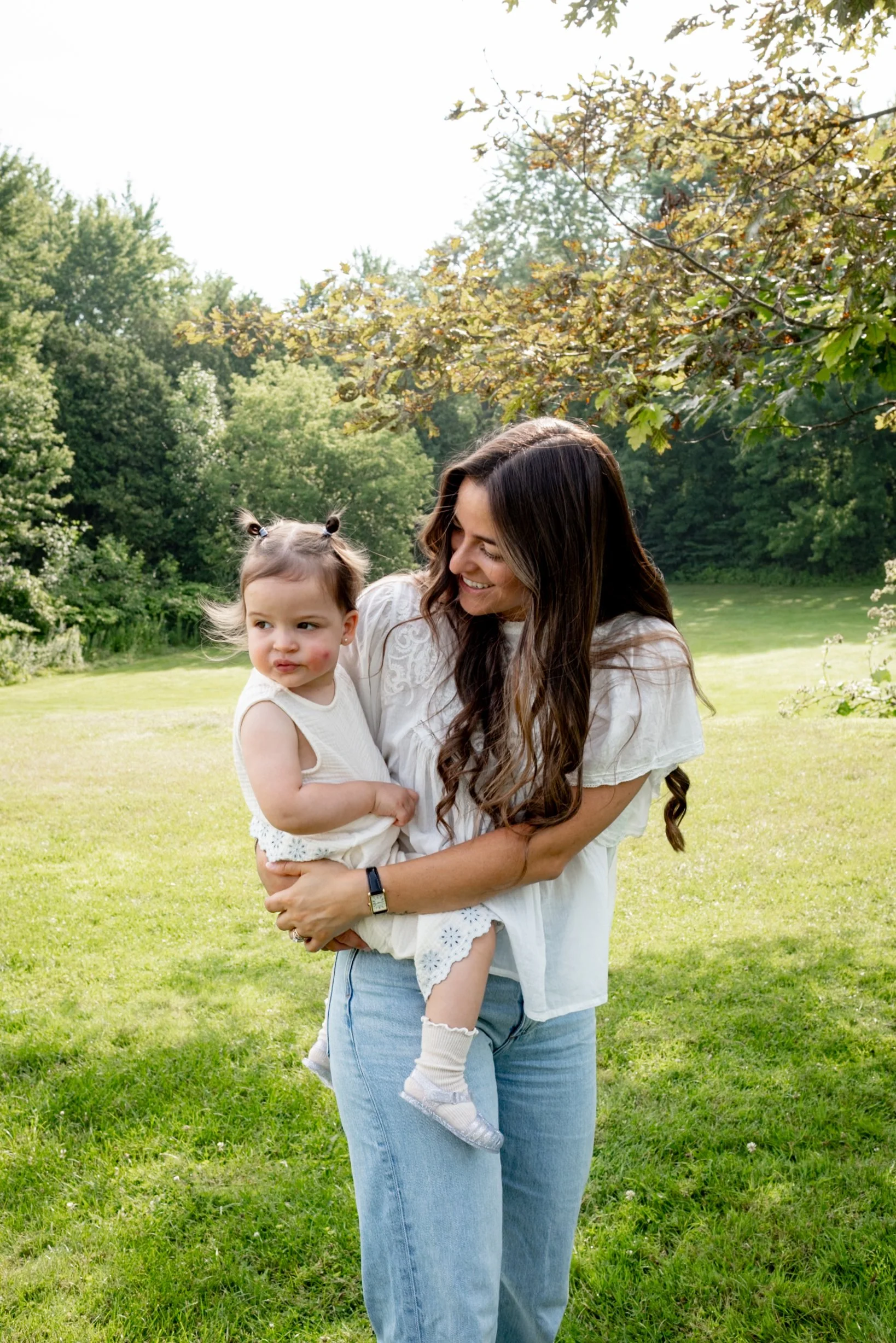 A woman with long brown hair, wearing a white blouse and blue jeans, holding a young girl with brown hair styled in pigtails, wearing a white dress, outside in a grassy area with trees in the background.
