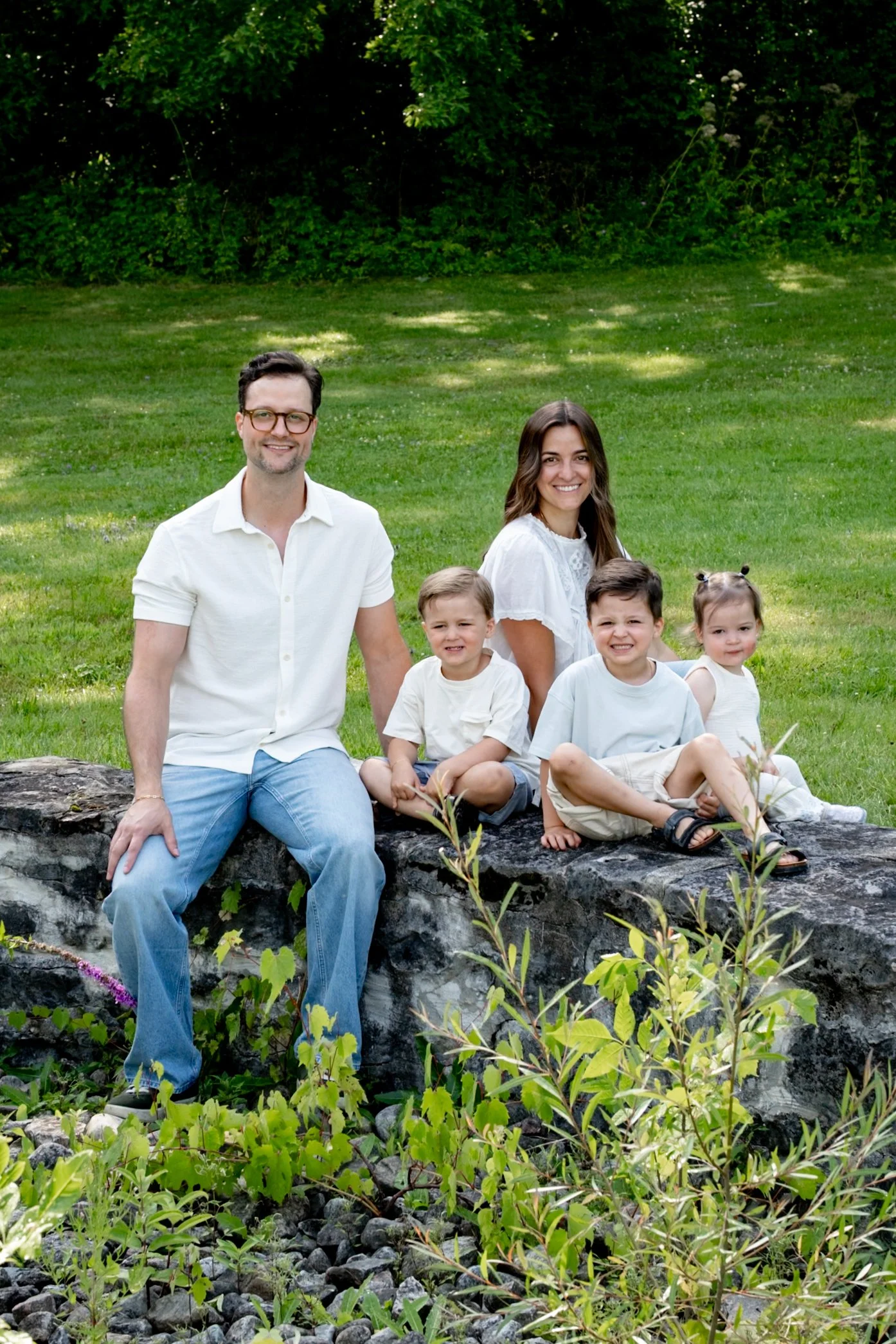 A family of six with three children, two women, and a man sit on a rock in a park with green grass and trees in the background. They are smiling and looking at the camera.
