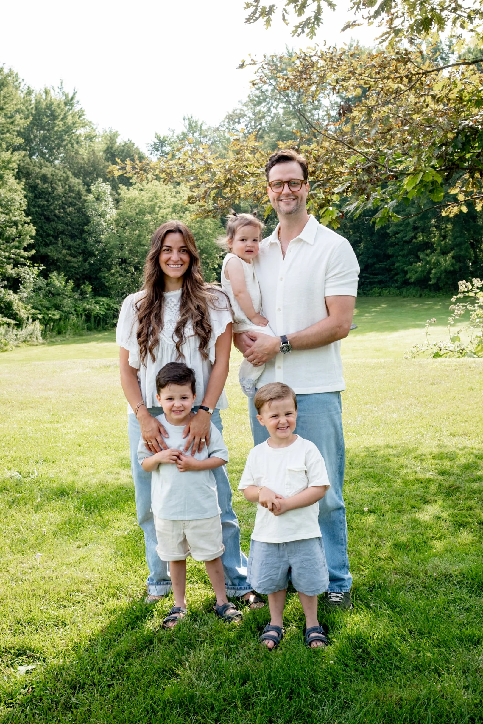 A family of six, including two adults and four children, standing outdoors on grass with trees in the background on a sunny day.