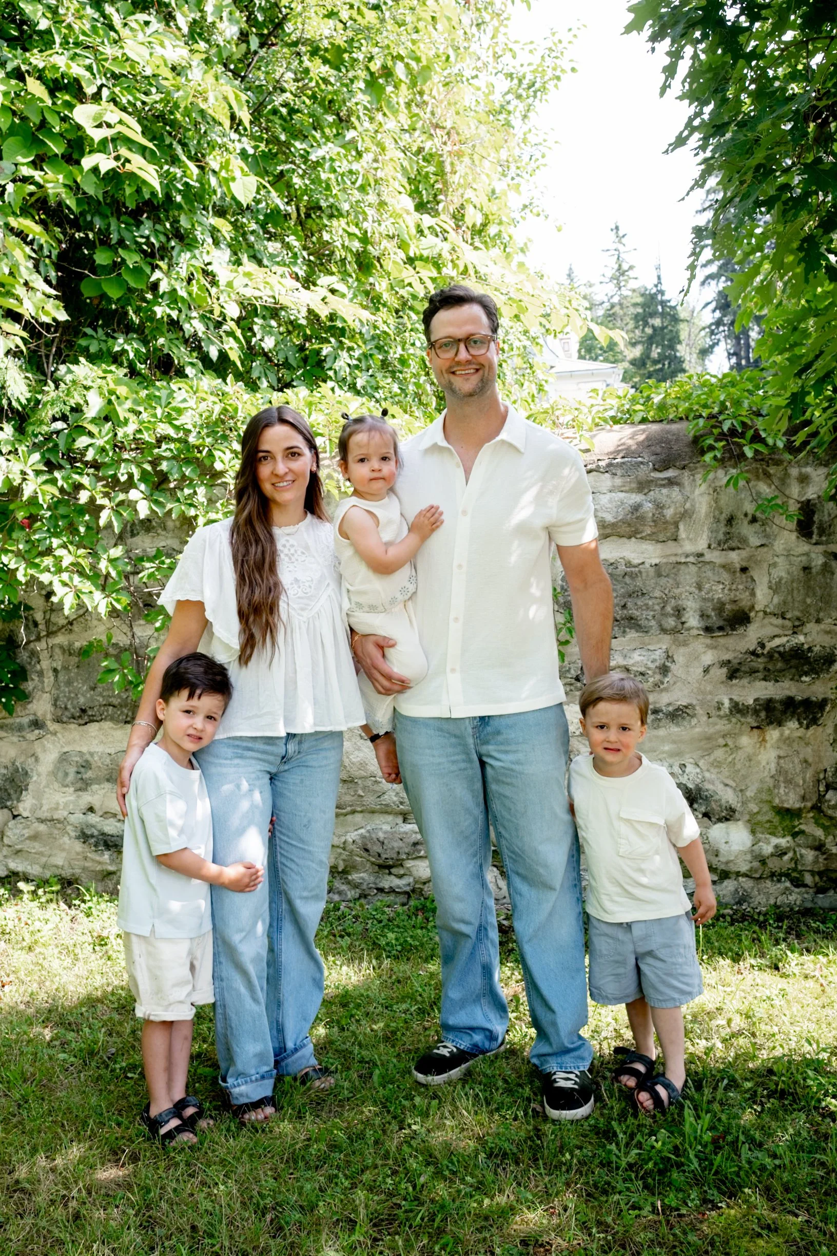 A family of seven standing outdoors in front of a stone wall and green foliage, smiling at the camera. The group includes two adults and five children, all dressed in light-colored clothing.