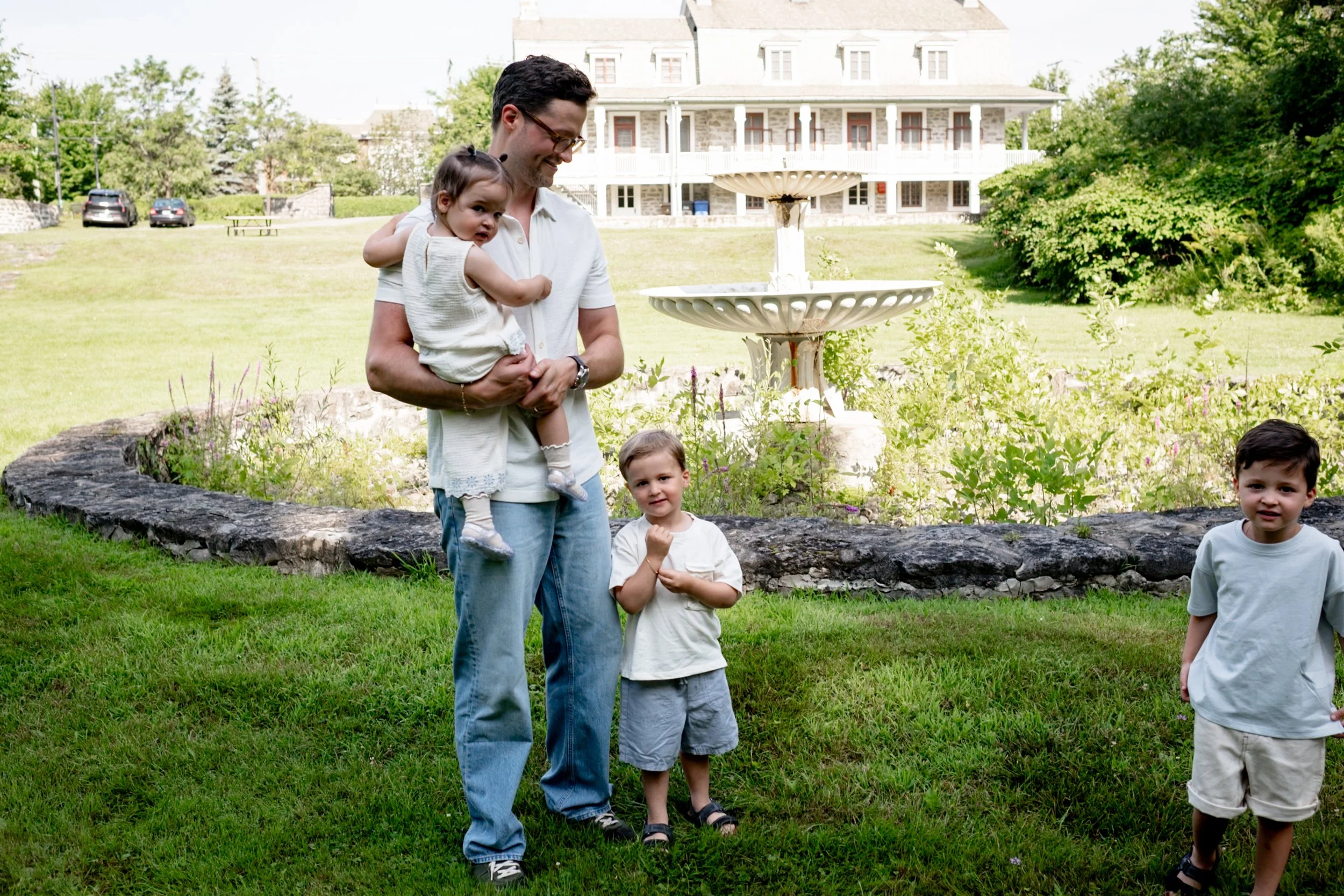 A man holding a young girl in a white dress, standing outdoors near a rock wall, with three young boys in casual clothing nearby, in front of a garden and a large house with a lawn and trees.