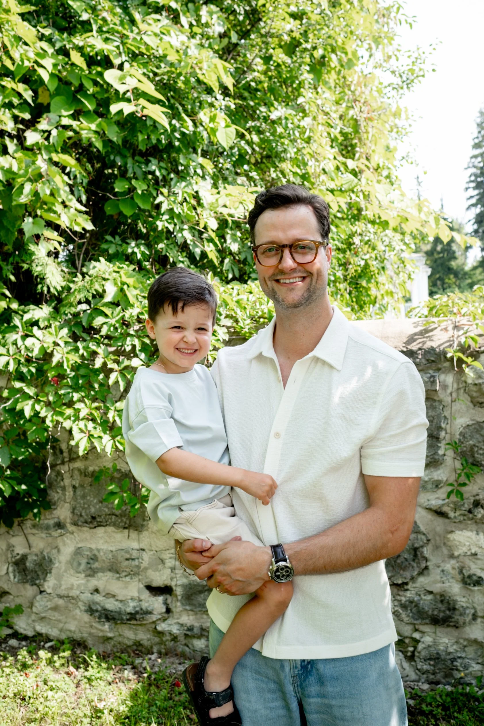 A man wearing glasses and a white polo shirt is holding a young boy in his arms in front of a lush green bush and a stone wall, both smiling happily towards the camera.
