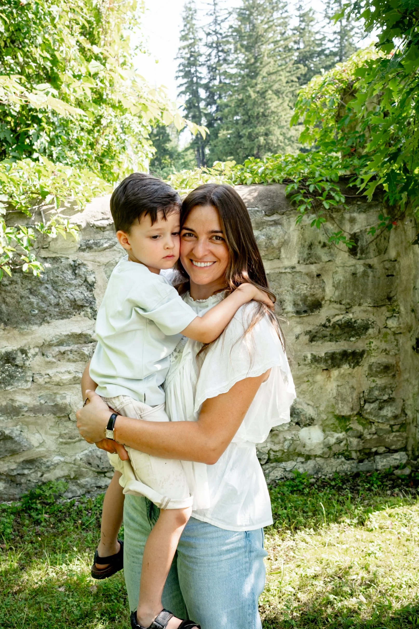 A smiling woman holding a young boy outdoors in front of a stone wall with greenery and trees in the background.
