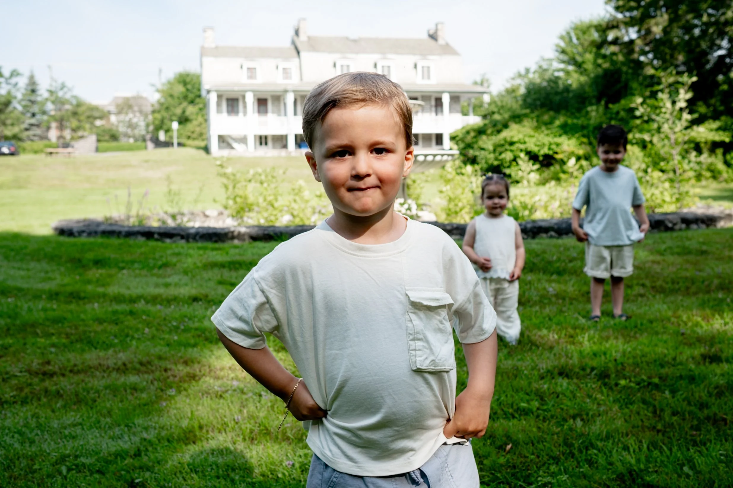 Young boy with light brown hair and a white t-shirt standing outdoors on a grassy lawn, with two other children and a large white house with a porch in the background.