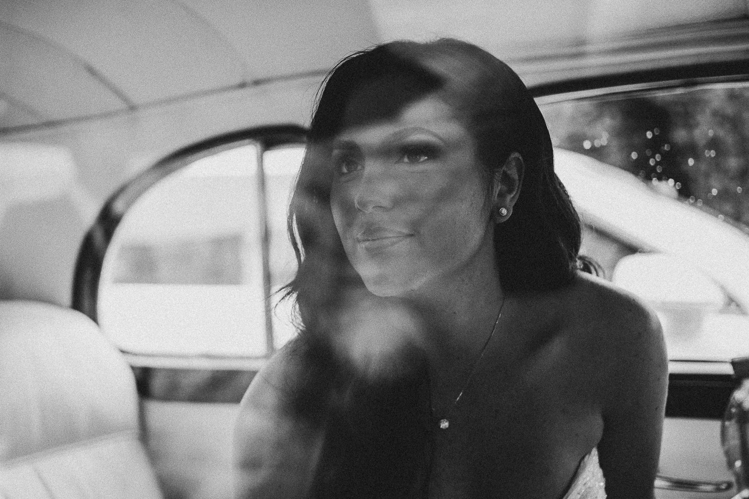 A black and white photo of a bride with dark hair, wearing earrings and a necklace, sitting inside a vintage car with a curved window, looking outside with a slight smile.