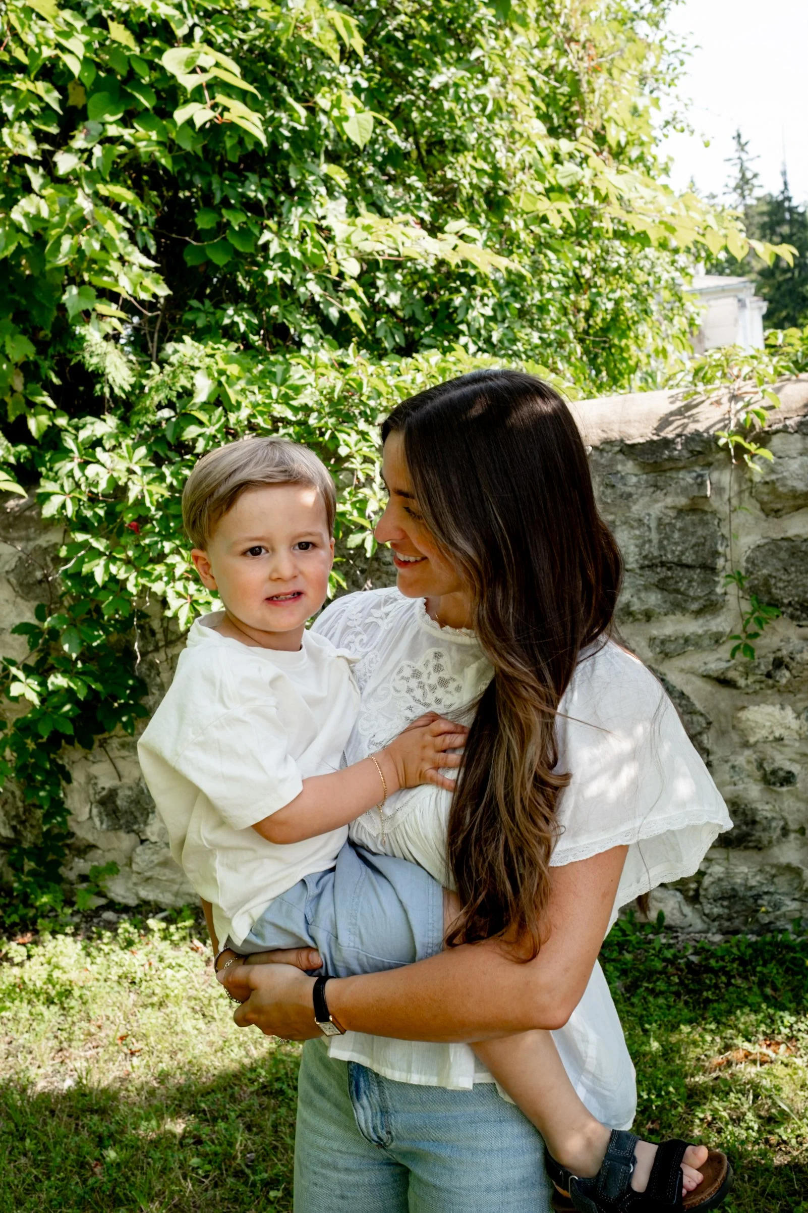 A woman with long brunette hair, smiling and holding a young boy in her arms outdoors. The boy has light brown hair and is wearing a white shirt and light blue shorts. They are standing in front of a bush with green leaves and a stone wall, with tree