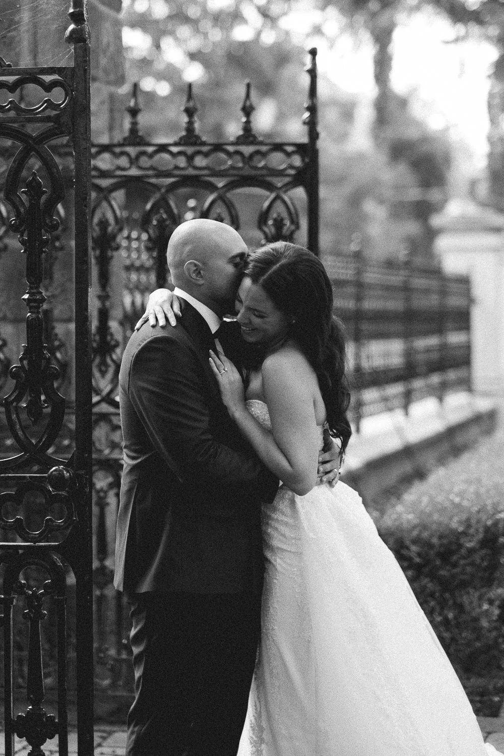 A newlywed couple sharing a kiss and hug outdoors near a decorative wrought iron gate, with a blurred background of trees and a fence in black and white.