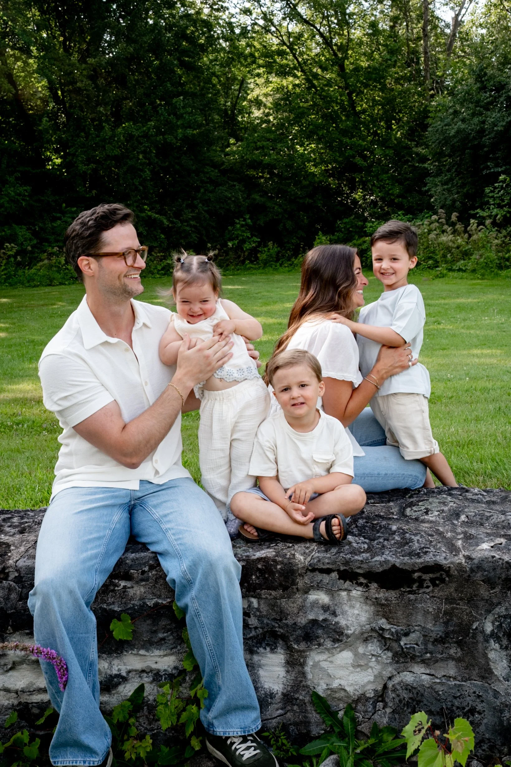 Family of five sitting outdoors on a large rock in a grassy park with trees in the background. The family includes a man, a woman, and three young children. The man is wearing glasses and a white shirt. The woman has long dark hair and is smiling whi