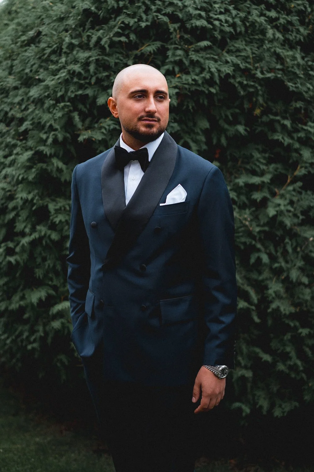 A groom in a dark blue tuxedo with a black satin shawl collar, white shirt, and black bow tie, standing outdoors in front of dense green bushes, looking to his left, with a watch on his left wrist.