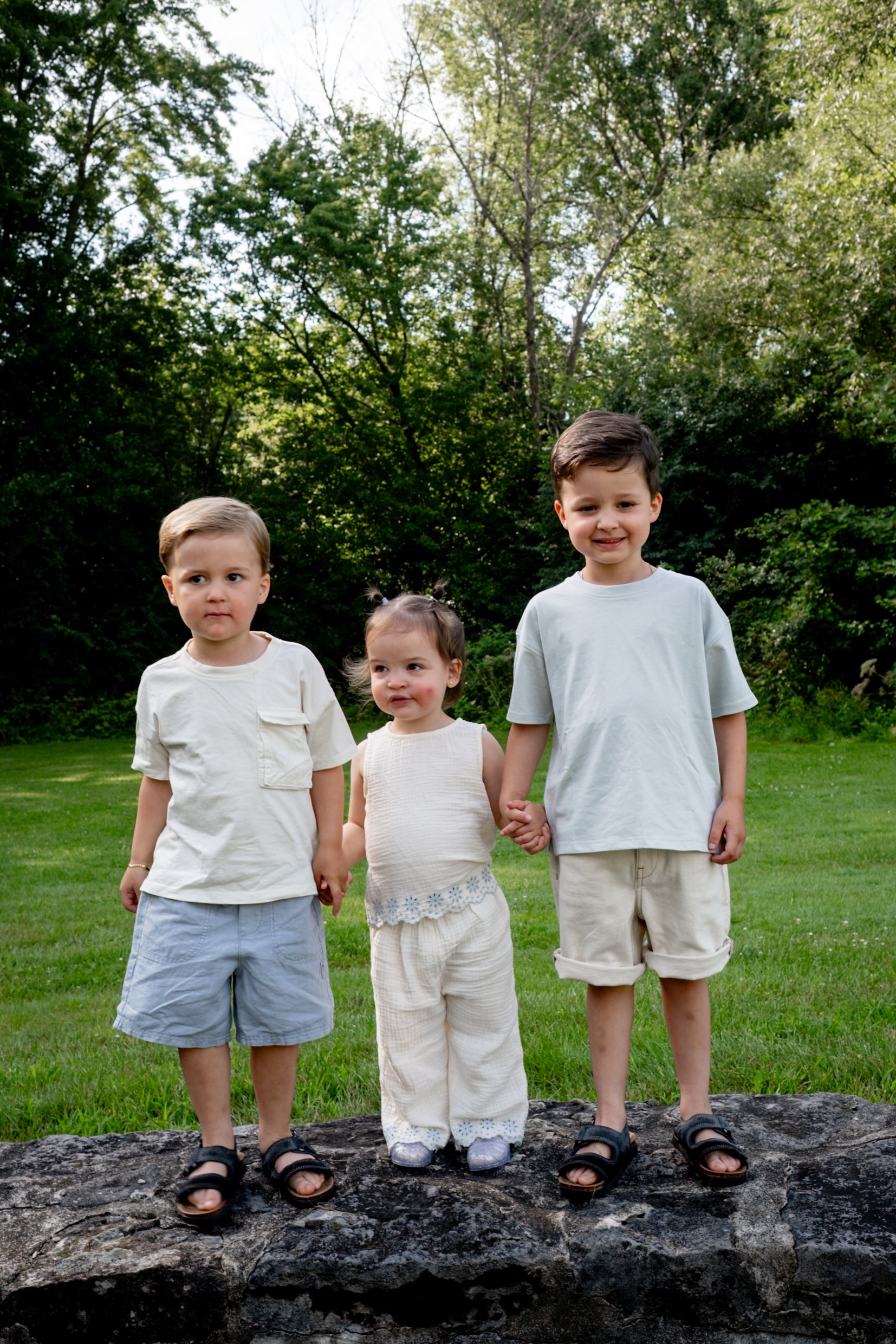 Four children, two boys and two girls, standing outdoors on a large fallen tree trunk in a park or forested area. They are holding hands and smiling or looking serious, with green trees and grass in the background.