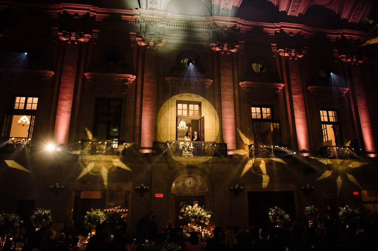 An elegant indoor event with two people on a balcony in front of large windows, illuminated by chandeliers and warm lighting, with a decorated dining area below and spotlight effects on the historical building facade.