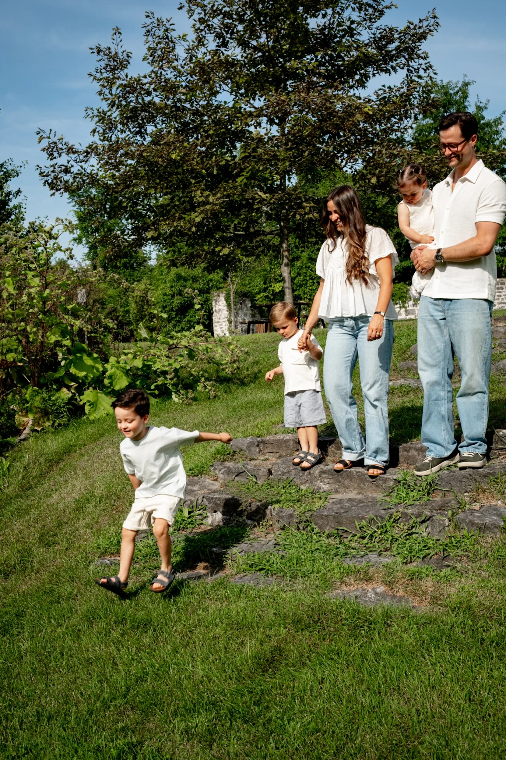 A happy family of five outdoors on a sunny day. The father holds a young girl, the mother holds a boy, and another boy is running down a stone staircase on green grass with trees and blue sky in the background.