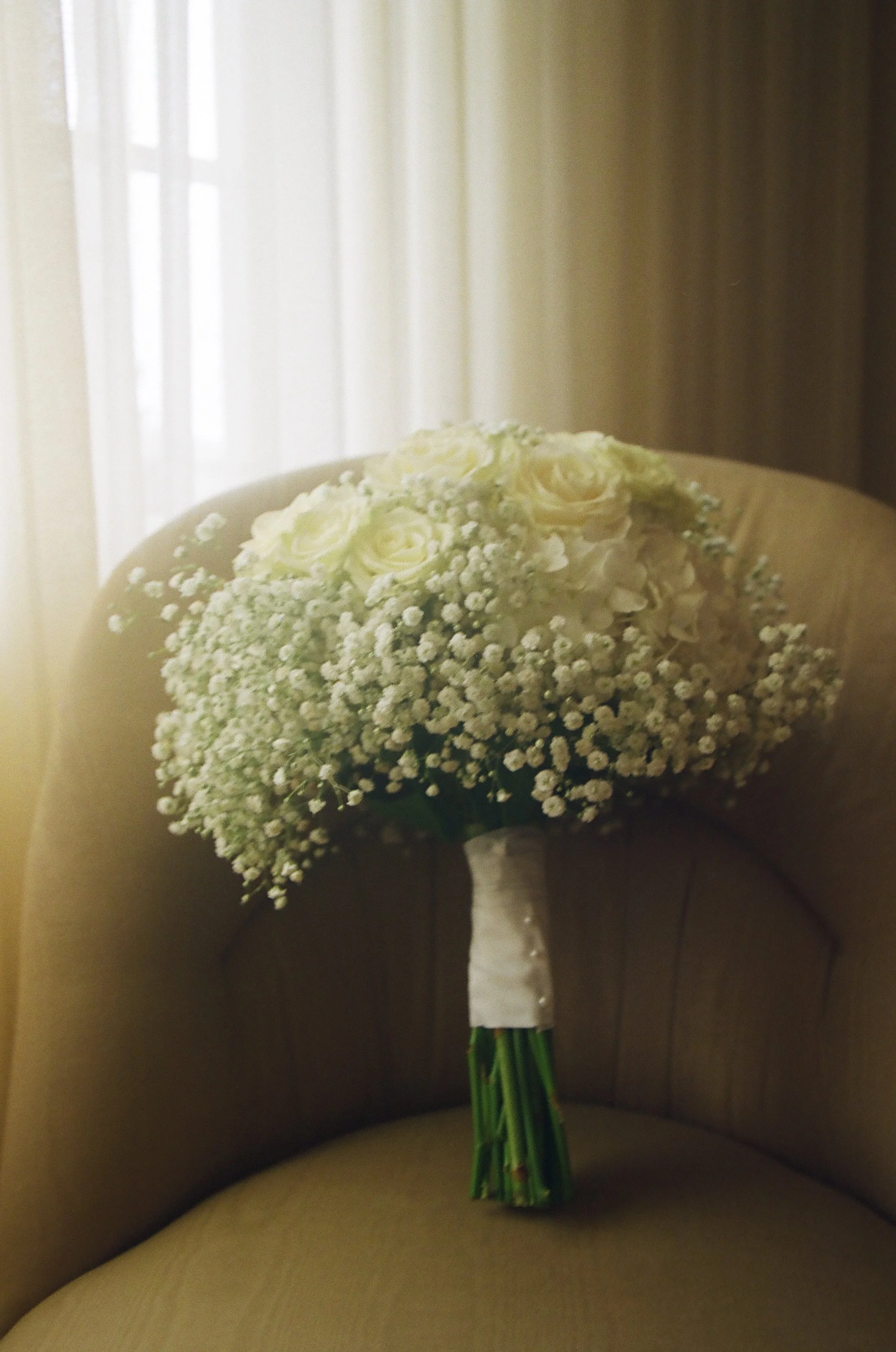 A white bridal bouquet with roses and baby's breath on a beige chair with a window in the background.