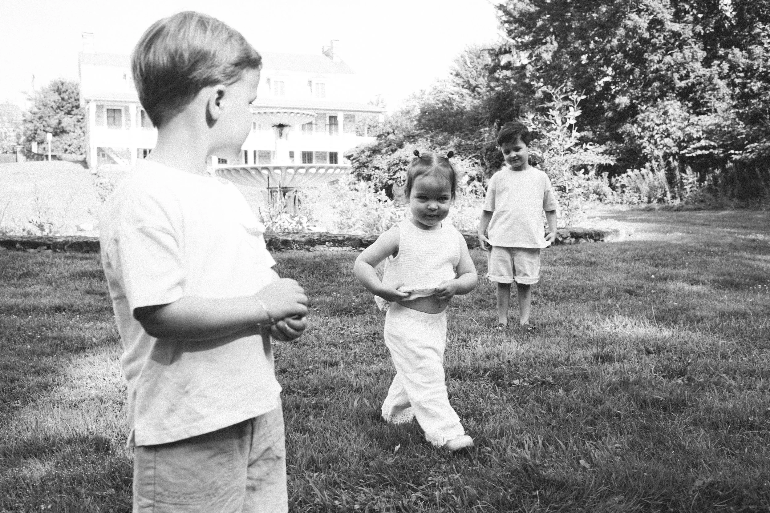 A black and white photo of three children playing outside on a grassy area with trees and a large house or hotel in the background. The children are dressed in casual clothes and seem to be enjoying their time.