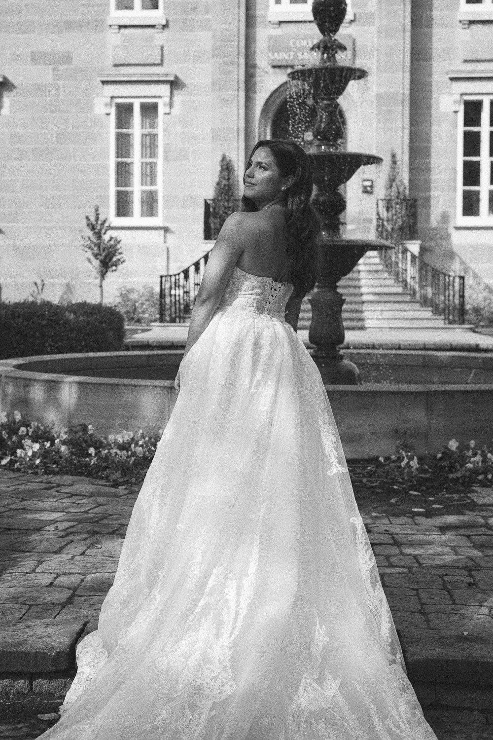A woman in a wedding dress standing outdoors near a fountain with a large building in the background.