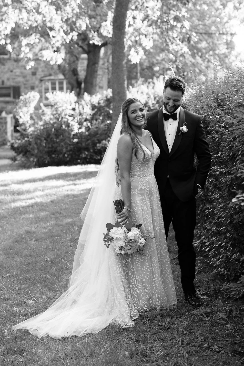 Black and white photo of a bride and groom smiling and walking outside in a garden, with trees and bushes in the background. The bride is holding a bouquet of flowers, and the groom is wearing a tuxedo with a bow tie.