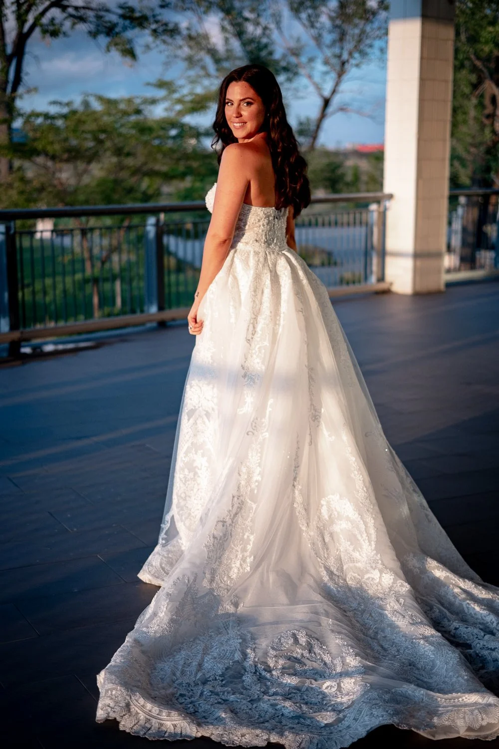 A bride in a white strapless wedding gown with lace details, standing outdoors on a balcony, smiling at the camera.