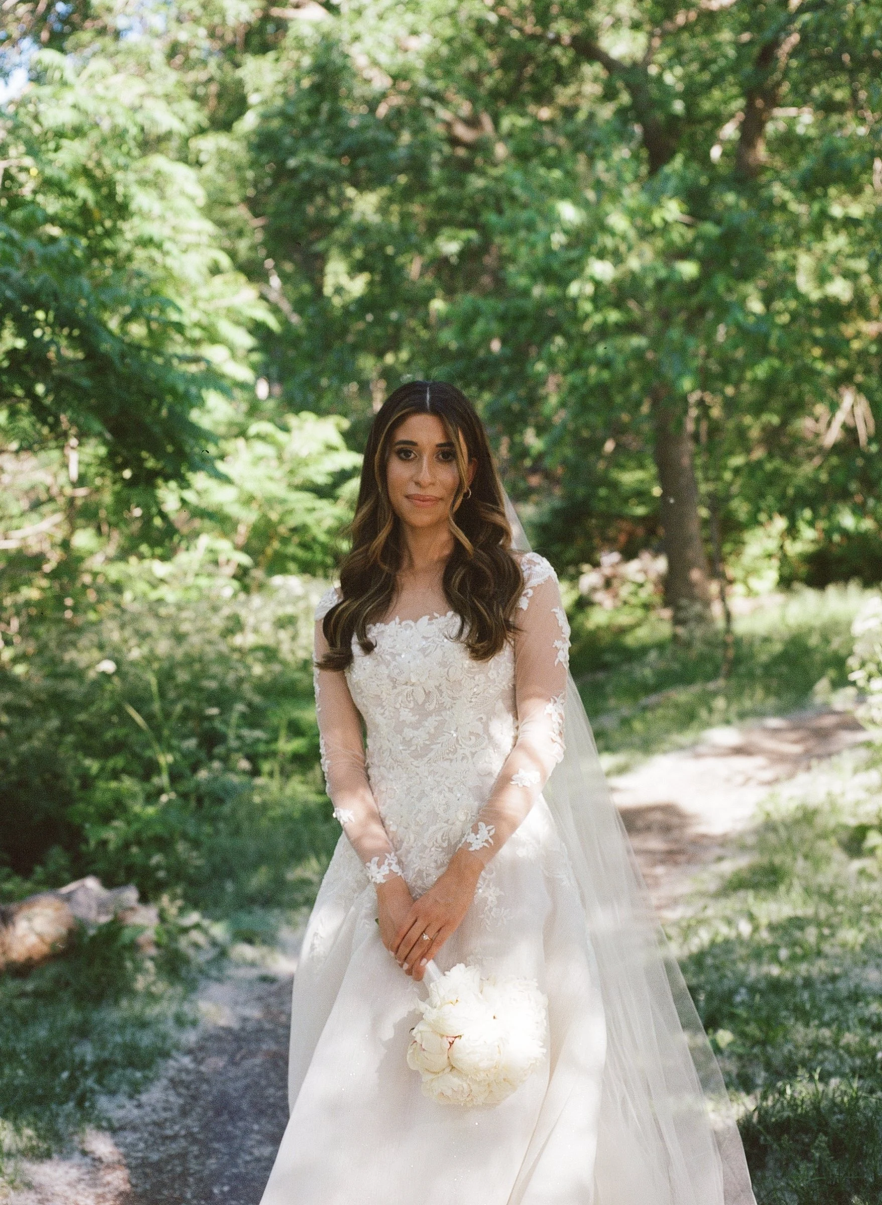 A bride standing on a wooded trail in a white wedding dress holding a bouquet of white flowers.