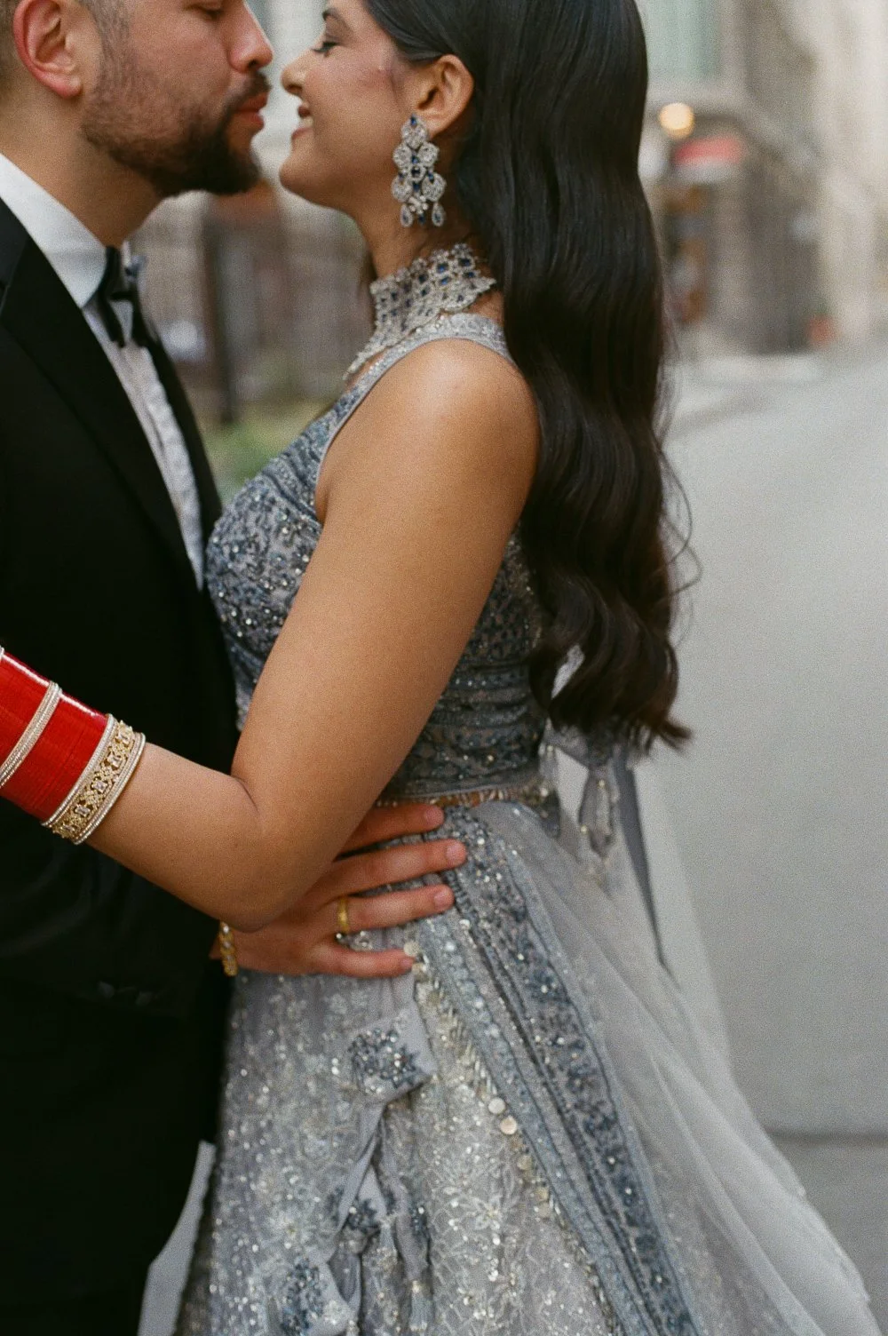 A couple in formal attire about to kiss, with the woman dressed in a silver embroidered gown and the man in a black suit, standing outdoors.