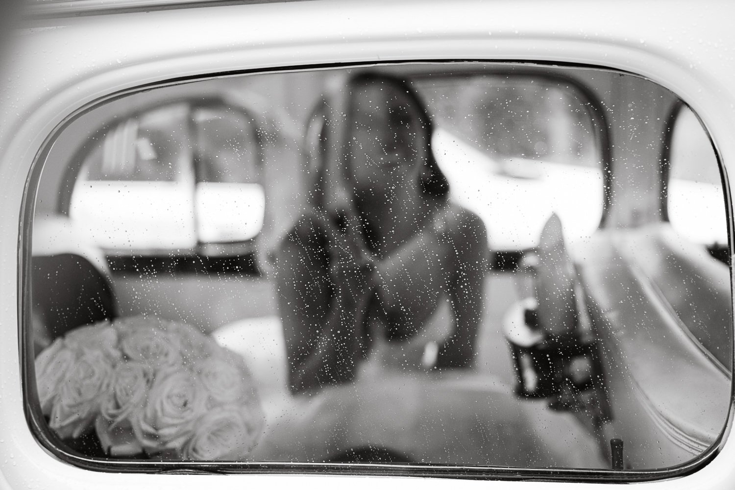 A bride in a car's rearview mirror, with rain droplets on the glass, and a bouquet of roses on the seat.