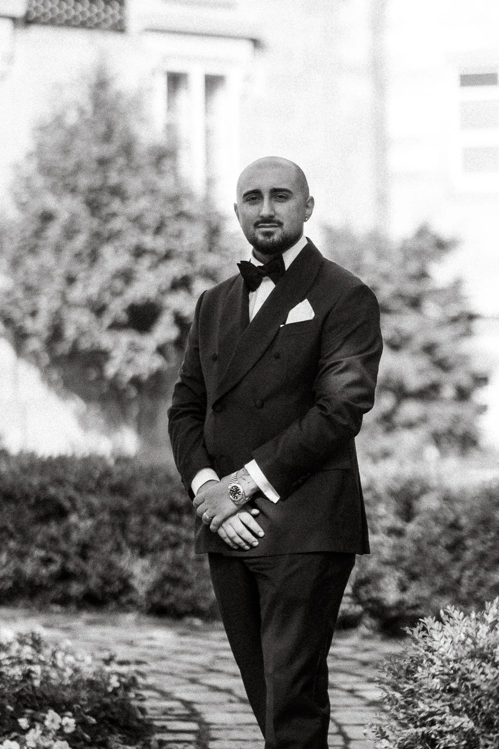 A man dressed in a tuxedo with a bow tie, standing outdoors with his hands clasped in front of him. The background features trees and buildings, and the photo is in black and white.