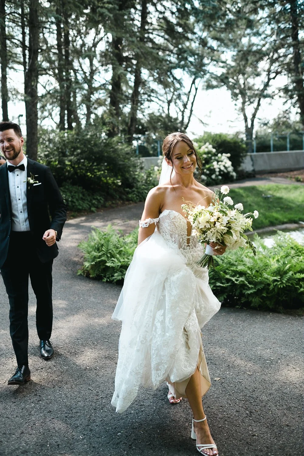 A bride in a white lace wedding gown with lace off-the-shoulder sleeves holding a bouquet of white flowers, smiling, walking outdoors on a paved path surrounded by greenery, with a groom in a black tuxedo walking beside her in the background.