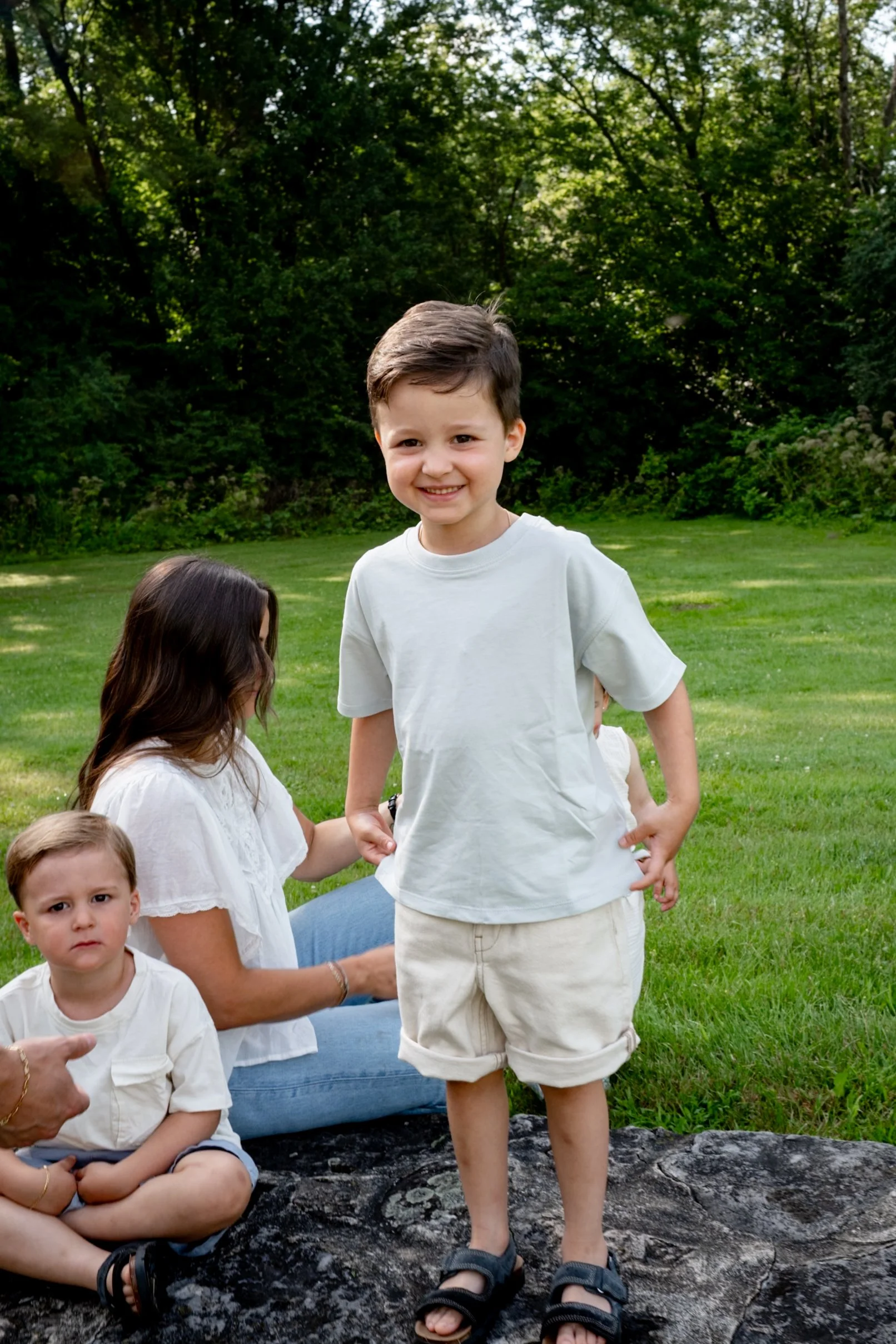 A group of children and an adult sitting on a large rock outdoors in a park with green grass and trees in the background. One boy is standing and smiling at the camera while others are seated or partially visible.