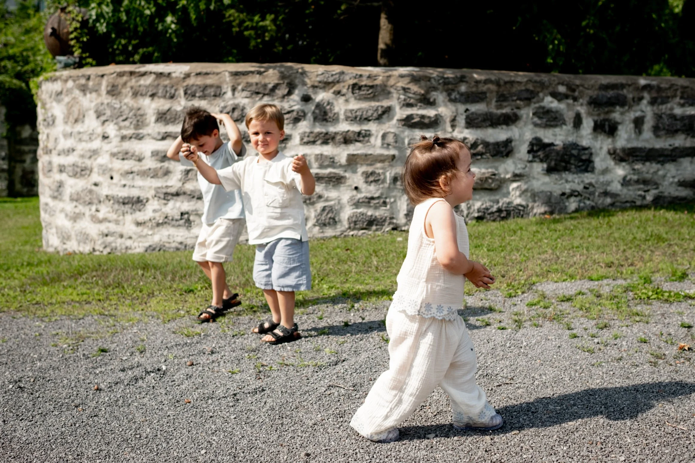 Three children playing outdoors near a stone wall. Two boys are standing together, one with his hand on his head, and the girl in front is walking away from the camera, wearing light-colored clothing.