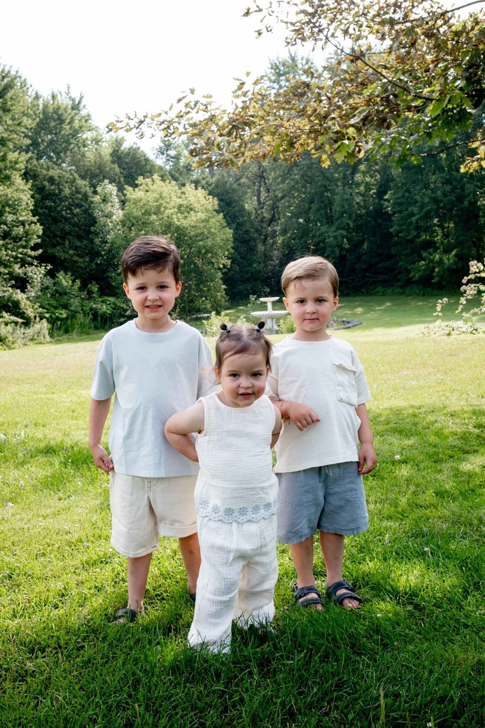 Four children standing on a lush green lawn with trees and a fountain in the background, outdoors on a sunny day.