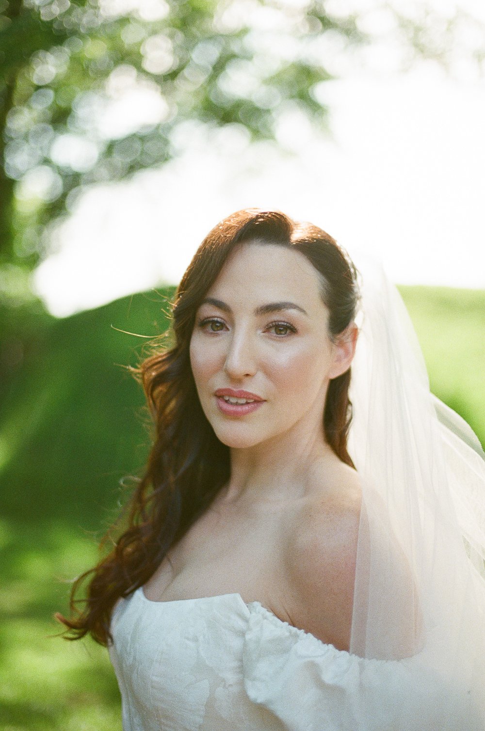 A woman in a white dress and veil outdoors, with sunlight filtering through trees in the background.