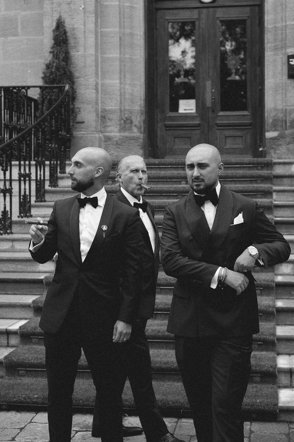 Groomsmen in tuxedos standing outside a building with stairs, two with cigars and one smoking. Black and white photograph.