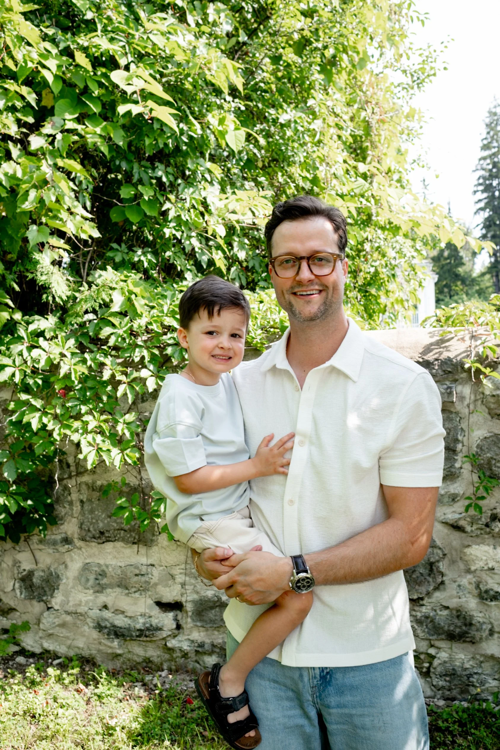 A man wearing glasses and a white polo shirt is holding a young boy, both smiling, outdoors with green bushes and trees in the background.