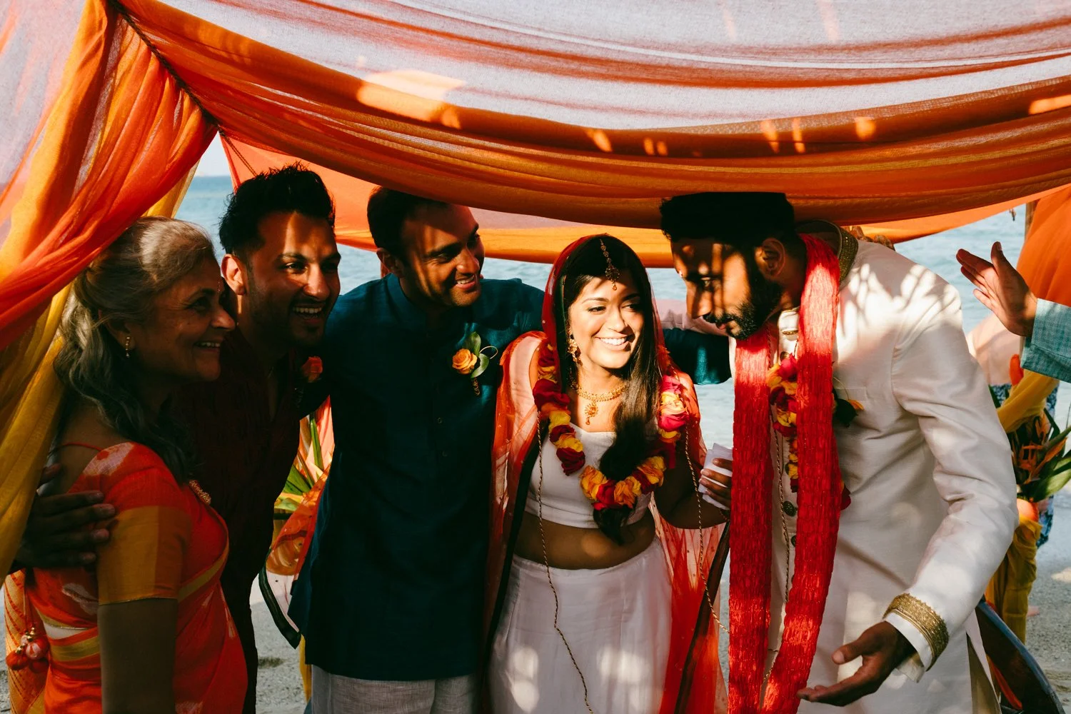 A group of diverse people in traditional Indian attire gathered under a colorful canopy near the beach, celebrating a wedding or cultural event.